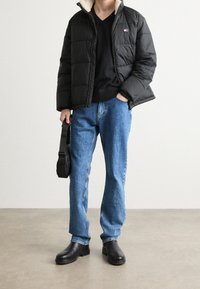 Doudoune noire avec un col en fausse fourrure, shirt noir à col en V, jean en denim bleu et bottes à talons noires. Tenant un sac à main noir.