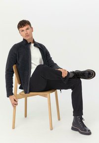 Veste en polaire gris foncé avec un col montant, associée à un t-shirt blanc et un pantalon noir. Modèle assis sur une chaise en bois clair, portant des bottes noires.