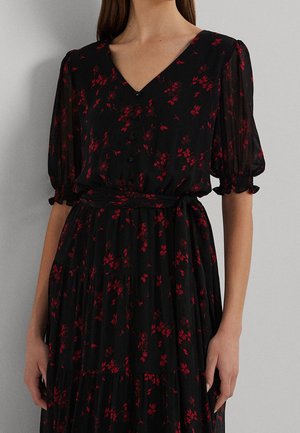 Frau trägt ein schwarzes Kleid mit rotem Blumenmuster, V-Ausschnitt, vorne geknöpft, gepuffte Ärmel bis zum Ellbogen und einen gebundenen Taillengürtel.