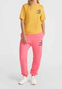 Camiseta de algodón amarilla con mangas cortas y texto en negro, combinada con pantalones deportivos rosas que presentan un texto similar y puños elásticos. Zapatillas blancas.