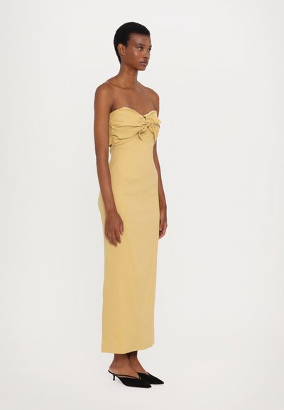 Gelbes, trägerloses Maxikleid mit knotenförmigem Vorderteil, figurbetonter Silhouette und strukturiertem Stoff. Schwarze spitze Schuhe runden das Outfit ab.