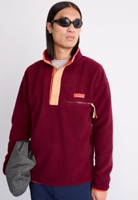 Bordo flis pulover s ovratnikom v oranžni in bež barvi, sprednjim gumbom, zadrgo, žepom in teksturirano tkanino. Model drži karirano torbo.
