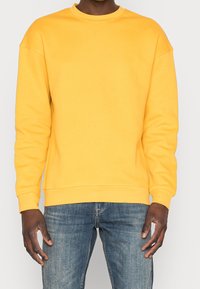 Gul crewneck sweatshirt i mjukt tyg, med avslappnad passform och ribbade muddar; bärs med blå jeans.