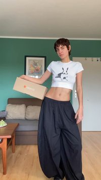 Biela skrátená tričko s potlačou čierneho zajaca, skombinované s voľnými čiernymi nohavicami. Model drží béžovú krabicu Zalando. Pozadie tvorí drevený stôl a zelená stena.