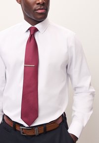 White dress shirt with a pink-patterned tie featuring a silver tie clip. Brown leather belt with a silver buckle, dark pants.