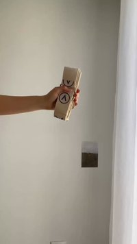 Χέρι κρατά διπλωμένη μπεζ υφασμάτινη ζώνη με κυκλικά μοβ λογότυπα μπροστά σε μια απλή τοίχο με ένα μικρό έργο τέχνης και κουρτίνα.