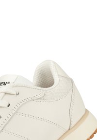 Detailní záběr na bílou tenisku, na které jsou vidět texturovaná pata, švy, tkaničky a polstrovaný límec na bílém pozadí.