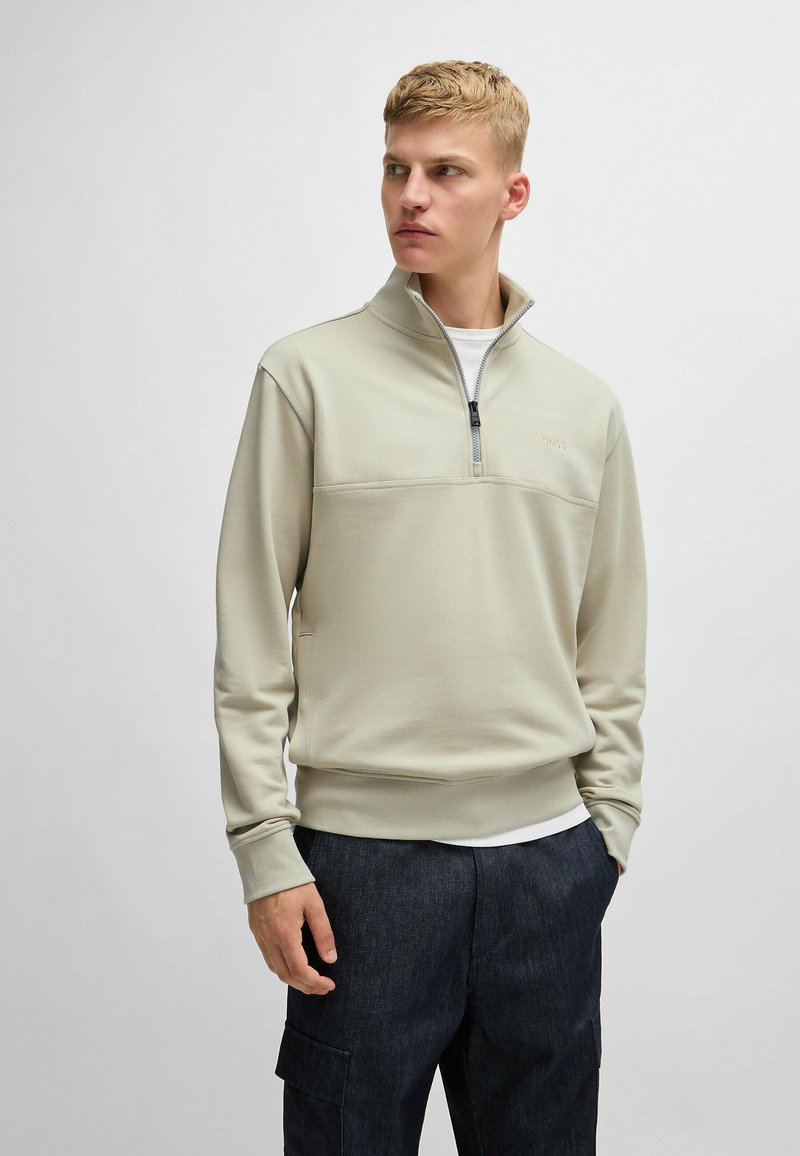 Felpa zip in beige chiaro con una sezione superiore texturizzata e polsini a costine. Abbinata a pantaloni cargo blu scuro, che mostrano un design casual.