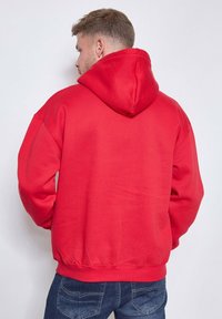 Homme aux cheveux courts portant un sweat à capuche rouge vif et un jean bleu, de dos contre un fond clair.