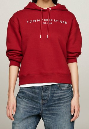 Personne portant un sweat à capuche rouge Tommy Hilfiger avec logo en texte et cordons de serrage, associé à un jean bleu, cadré à mi-torse et bras.