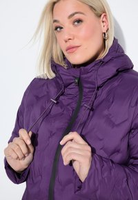 Chaqueta de plumas púrpura con capucha, que cuenta con cordones y un cierre frontal de cremallera. La tela tiene una textura suave y un diseño ligero.