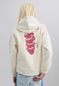 Persona con cabello rubio largo que lleva una sudadera color blanco roto con un gráfico de fresas rojas y texto parcial en la parte de atrás, combinada con jeans de color azul claro.