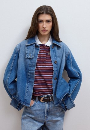 Giovane donna con lunghi capelli castani che indossa una giacca di jeans blu, una polo a righe bordeaux e jeans blu, in piedi con le mani nelle tasche.