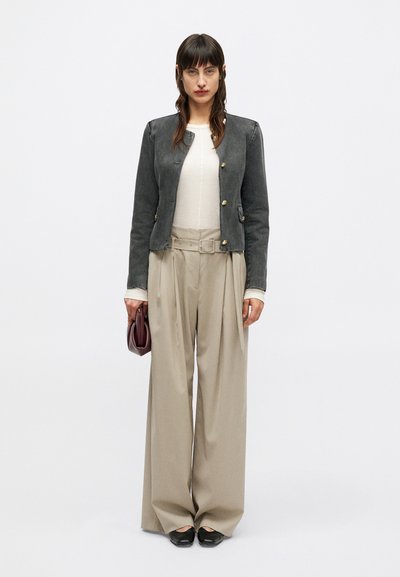 Γυναίκα που φοράει ένα εφαρμοστό σκούρο γκρι σακάκι με χρυσά κουμπιά, κρέμ μπλούζα, ψηλόμεσα φαρδιά μπεζ παντελόνια, μαύρα παπούτσια, κρατώντας μια μικρή μπορντό τσάντα.