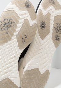 Běžecké boty s texturovanou gumovou podrážkou ve světle béžové a tmavě šedé barvě, zdobené květinovými vzory a vlnitým designem na spodní straně.