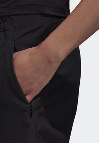 Pantalon de sport noir à la texture lisse, doté d'une poche latérale et d'une ceinture élastique, partiellement visible avec une main dans la poche.