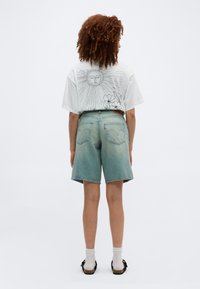 Γυναίκα με σγουρά μαλλιά που φοράει λευκό μπλουζάκι με σχέδιο ήλιου και λουλούδι, ανοιχτό μπλε τζιν σορτς, λευκές κάλτσες και μαύρα σανδάλια, στέκεται με την πλάτη προς το μέρος μας.