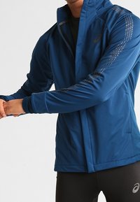 Persona che indossa una giacca blu con zip dalle maniche fantasiose e leggings neri da allenamento, mentre allunga le braccia attraverso il corpo.