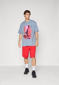 Ung mand iført blå grafisk t-shirt, røde shorts, hvide Nike sokker og sorte sneakers, der står foran en ensfarvet grå baggrund.