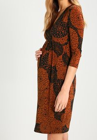 Raskauden mekko ruskeassa ja mustassa kukkakuosissa, jossa on kolme neljännesosaa hihasta, v-pääntie ja kapeasti mukautuva bodice, jossa on gathered-tyyli.