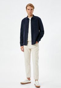 Camisa de botones azul marino con un bolsillo, suéter color crema claro, pantalones beige y zapatillas blancas con suelas marrones, de pie sobre un fondo neutro.