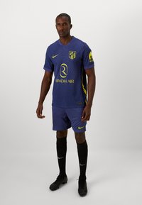 Voetbaltenue met een paarse shirt met verticale strepen, gele accenten en een logo, gecombineerd met bijpassende shorts en zwarte sokken.