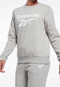 Grå sweatshirt tillverkad av bomullsblandat material, med en vit Reebok-logotyp fram, ribbad rund hals och elastisk midja.