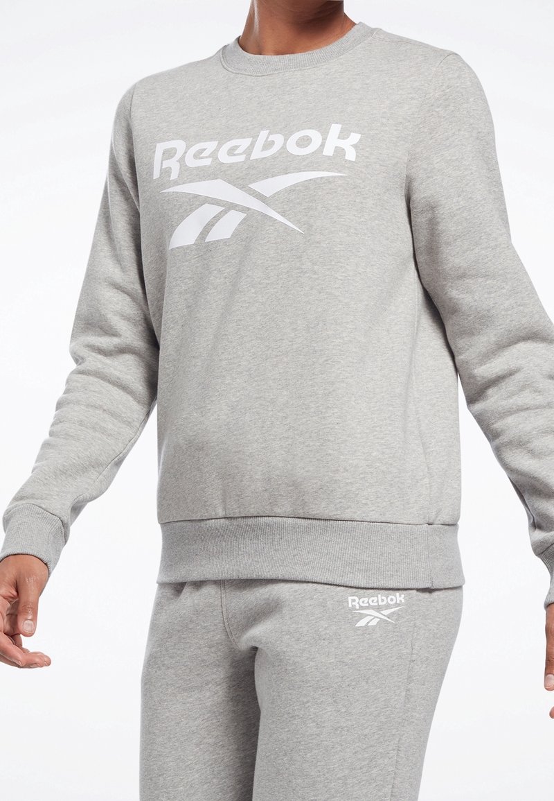 Grå sweatshirt tillverkad av bomullsblandat material, med en vit Reebok-logotyp fram, ribbad rund hals och elastisk midja.