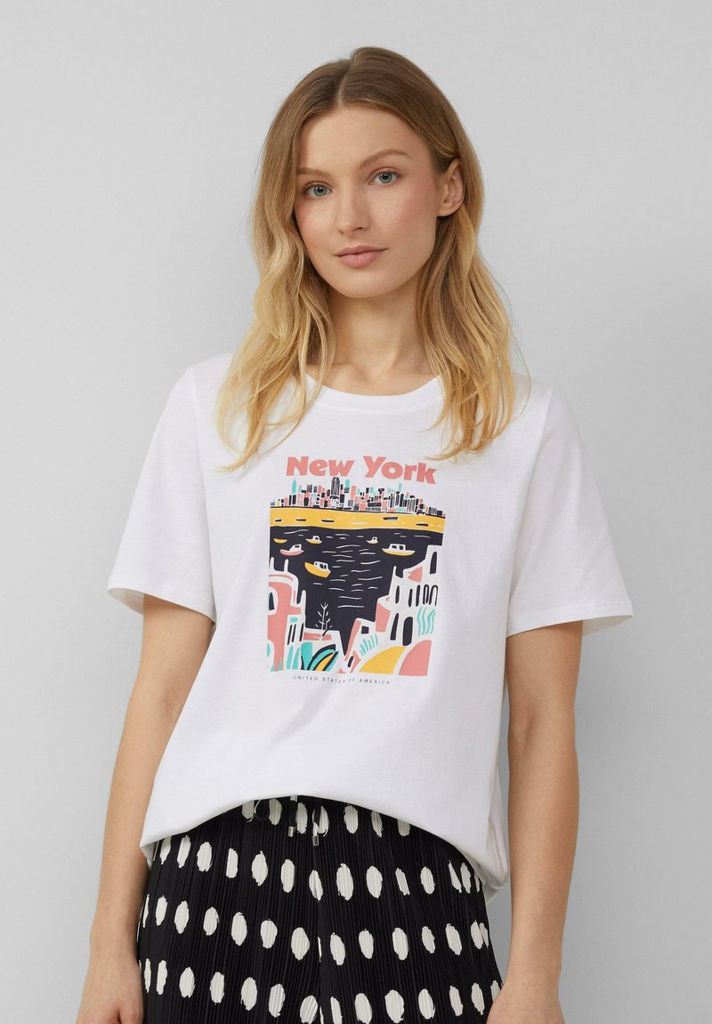 T-shirt en coton blanc arborant un graphique coloré représentant un paysage urbain de New York avec des bateaux sur l'eau, le tout sur un fond abstrait vibrant.