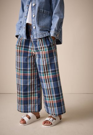 Person iført blå, grøn og rød ternet vidde bukser, en lyseblå denimjakke over en hvid top og hvide sandaler med spænde.