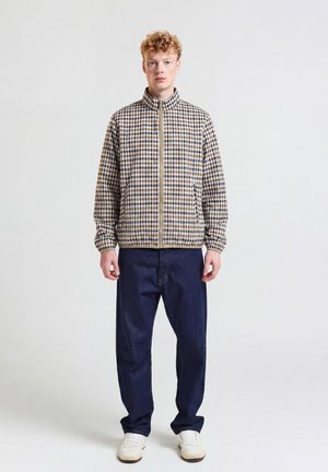Junger Mann mit lockigem roten Haar, der eine beige und marineblaue karierte Reißverschlussjacke, lockere dunkelblaue Jeans und weiße Sneakers trägt, steht vor weißem Hintergrund.