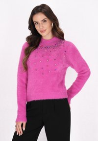 Uldent lyserød sweater med lange ærmer, der har farverige juvel-accenter rundt om halsudskæringen. Bæres med sorte bukser.