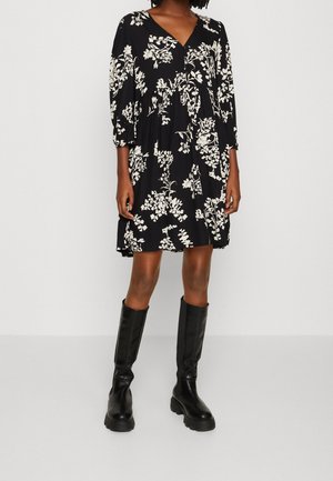 Schwarzes Blumenkleid mit V-Ausschnitt und drei Viertelärmeln, mit geraffter Taille und fließendem Rock. Kombiniert mit knielangen schwarzen Stiefeln.
