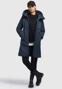 Marineblau wasserfester Parka mit Kapuze, ausgestattet mit einem Frontreißverschluss, seitlichen Taschen, kombiniert mit einem schwarzen Pullover, schwarzen Hosen und schwarzen Stiefeln.