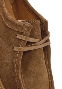 Close-up van een bruine suède schoen met gedetailleerde stiksels en een voorzijde met veters, zachte lederen textuur en een opgevouwen tong.