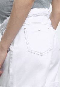 Άτομο που φοράει λευκά τζιν σορτς με ψηλή μέση και γκρι αμάνικο τοπ, με το ένα χέρι να ακουμπάει στο πλάι.