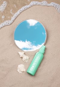 Botella de spray verde menta etiquetada "IDAWARG" al lado de un espejo redondo que refleja nubes, colocada en una playa de arena con dos conchas blancas.