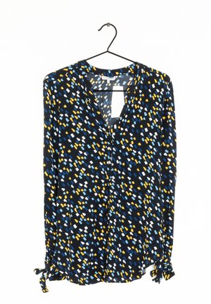 Blouse noire à manches longues avec un motif abstrait de points bleu, blanc et jaune, fermeture à boutons sur le devant et poignets noués, suspendue sur un cintre noir.