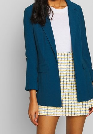 Nainen, jolla on päällään valkoisen topin päällä turkoosivihreä bleiseri ja keltainen ja sininen ruudullinen minihame, seisoo yksiväristä taustaa vasten.