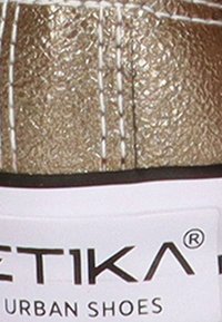 Zapatilla texturizada en dorado con costuras blancas, que cuenta con un acolchado en el tobillo y una suela negra. La etiqueta dice "ETIKA URBAN SHOES."