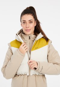 Bicolora pūkinė striukė, turinti bežę ir baltą dizainą su geltonu akcentu ant pečių, elastingomis rankovėmis ir raišteliu juosmens srityje.