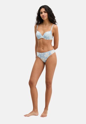 Frau steht barfuß, trägt einen hellblauen BH mit Blumenmuster und passende Bikini-Unterwäsche vor einem schlichten weißen Hintergrund.