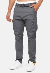 Pantaloni cargo in cotone grigio, con più tasche laterali, un design aderente e una cintura nera con fibbia in metallo. Sneakers bianche completano l'outfit.