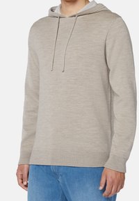 Ljusgrå huvtröja i ett mjukt, texturerat tyg. Har en dragkordshuva och ribbade ärmslut. Bärs över blå jeans.