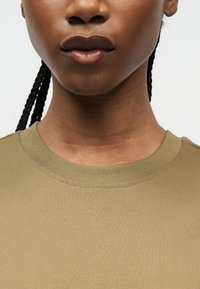 Κοντινό πλάνο στο κάτω μέρος του προσώπου και τον λαιμό ενός ατόμου με πλεγμένα μαλλιά, που φοράει απλή μπλούζα με στρογγυλή λαιμόκοψη σε ελιά πράσινο χρώμα, με φόντο ένα λευκό τοπίο.