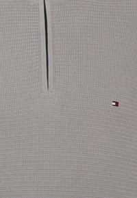 Pullover lavorato a maglia grigio chiaro con zip a quarto, caratterizzato da un motivo testurizzato e un piccolo logo in rosso, bianco e blu vicino all'orlo.