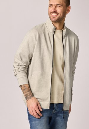 Hombre sonriendo y mirando hacia la derecha, vistiendo una chaqueta ligera beige con cremallera sobre una camisa beige y vaqueros azules, con un tatuaje visible en el antebrazo izquierdo.