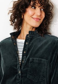 Chemise en velours côtelé vert foncé avec col montant, boutons sur le devant et poche poitrine. En dessous, un haut à rayures blanches et bleues est visible.