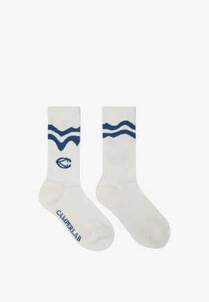 Calzini da uomo bianchi con trama a coste, motivi a onde blu scuro vicino alla parte superiore, un logo circolare e la scritta "CAMPERLAB" sul piede.