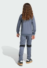 Chándal atlético gris con tejido texturizado, con detalles negros en los pantalones. Niño que lleva zapatos deportivos con tres rayas y suela de goma.